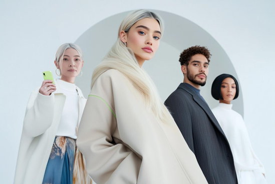 Four fashion models pose in a minimalist studio with a light gray arch backdrop. The central figure wears an oversized cream-colored coat, while three others stand behind wearing modern, neutral-toned outfits. One person holds a bright green smartphone, adding a pop of color to the contemporary fashion scene.