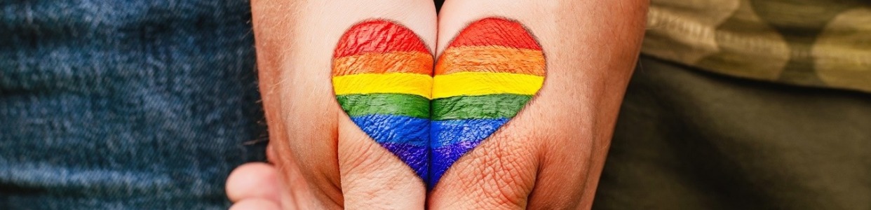 Two hands are placed side by side with a rainbow-striped heart painted across them. The heart features the colors red, orange, yellow, green, blue, and purple, representing the LGBTQ+ pride flag.