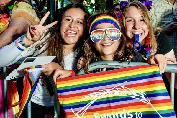 Three people at a Pride event smile and pose behind a metal barrier. One flashes a peace sign, another wears rainbow sunglasses and a headband, and the third blows a whistle. They hold a rainbow-striped banner with the text "Swords to..." and a white graphic of a sword transforming into a wheat stalk.